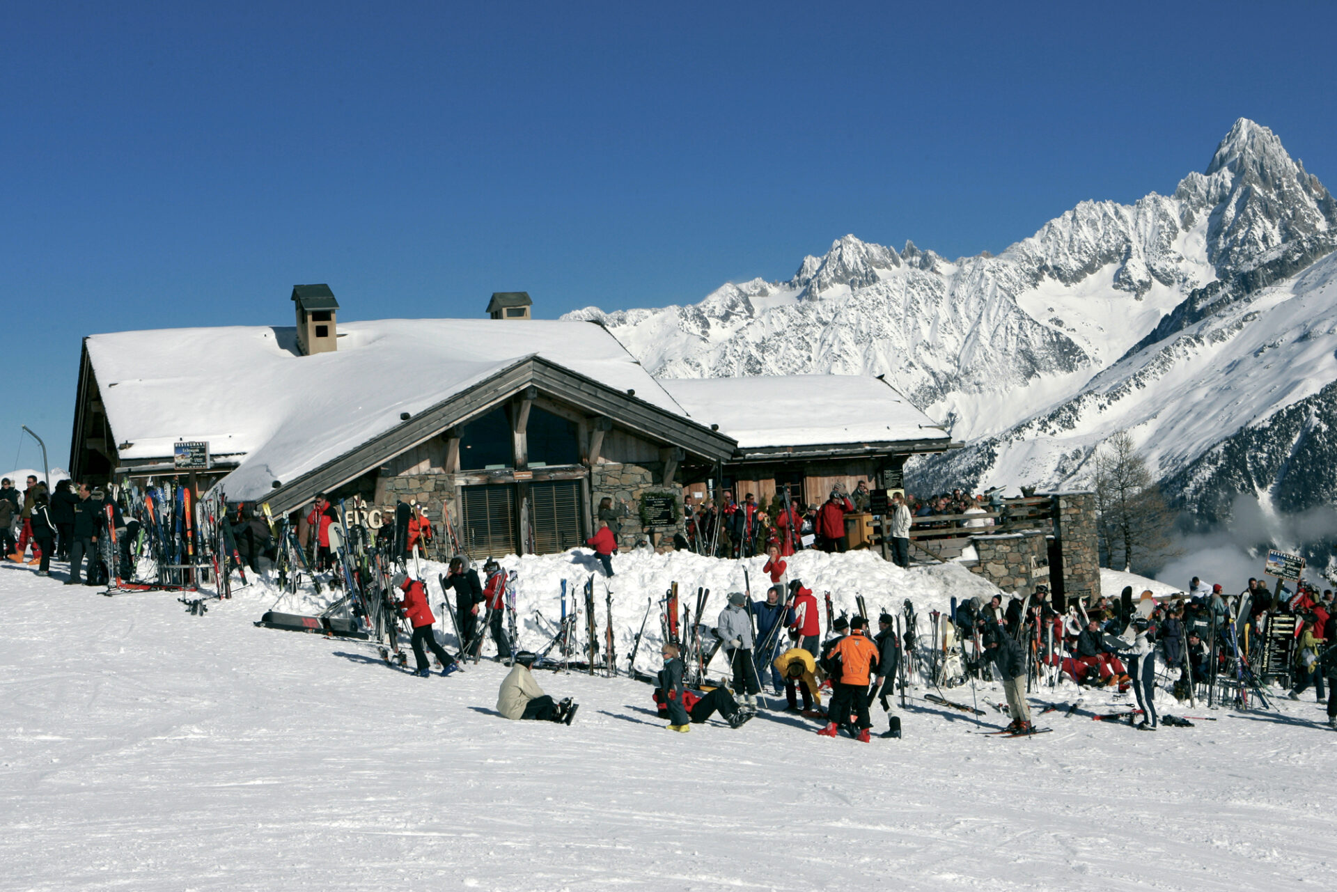 A restaurant on the slopes in Chamonix. There are lots of skis lined up outside and the roof of the restaurant is covered in snow.