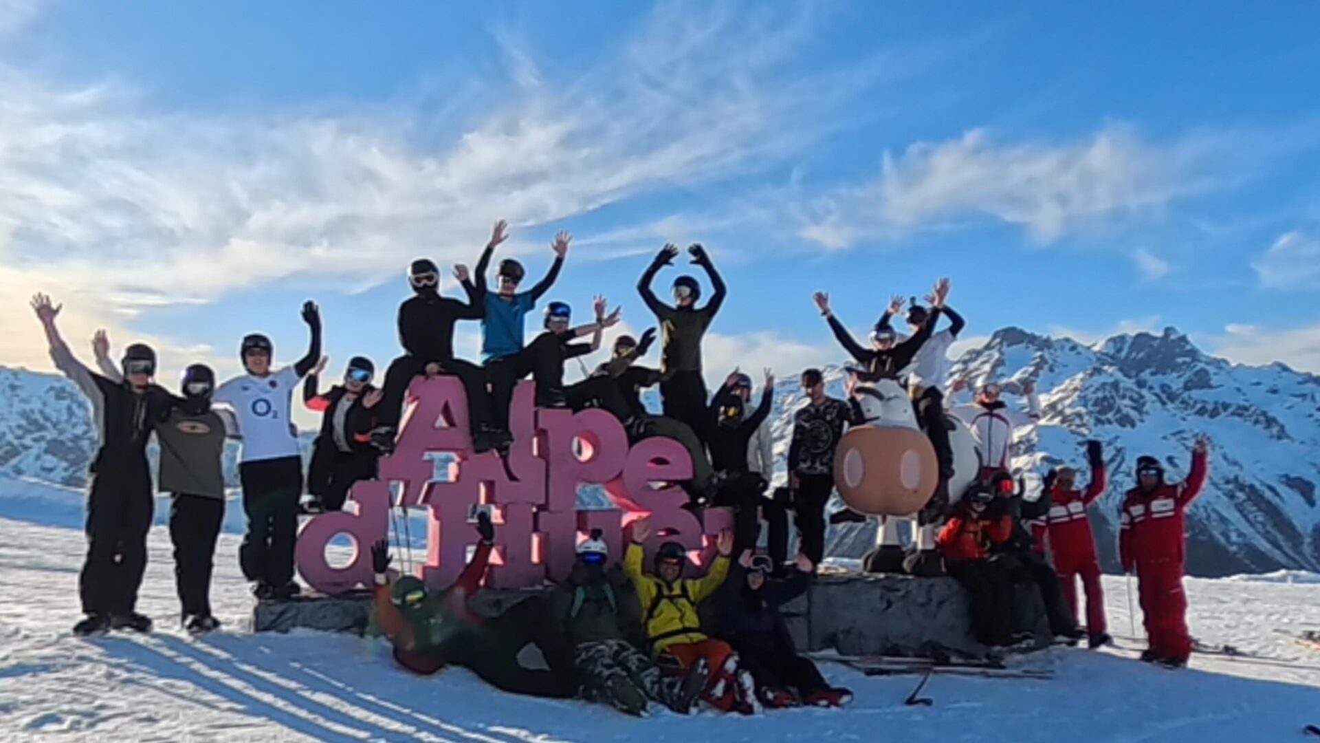 A big group of students with their hands in the air, sitting on the Alpe d'Huez sign